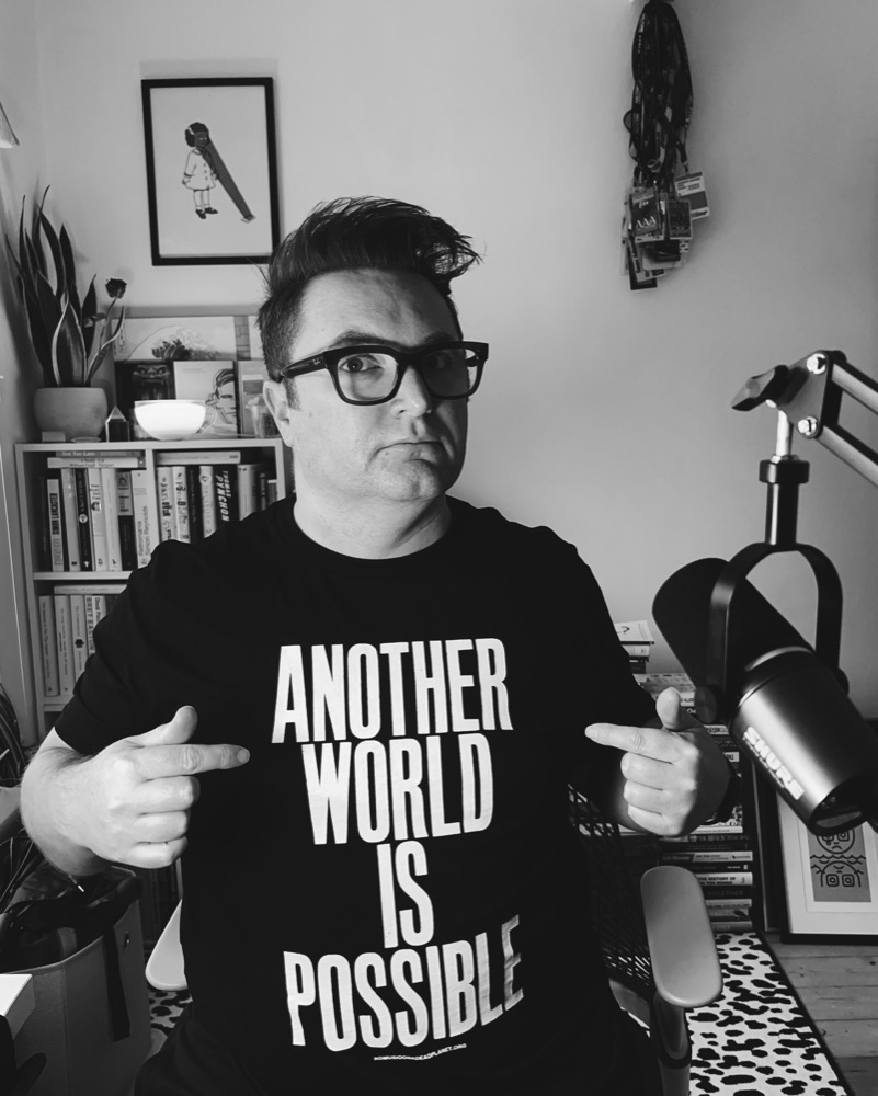 Sean Adams in his home studio, pointing at his t-shirt which reads 'Another World Is Possible'